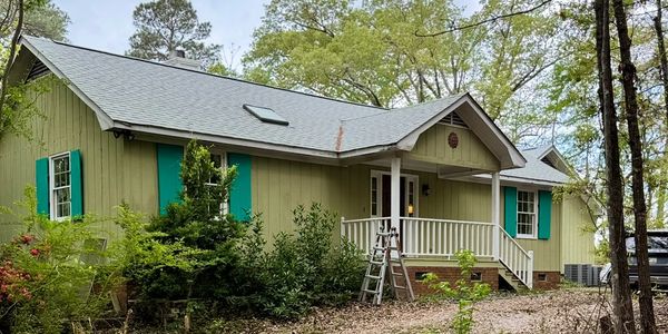 A green house with teal shutters surrounded by trees.