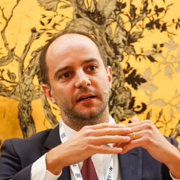 Man in suit speaking with hands clasped against a decorative backdrop.