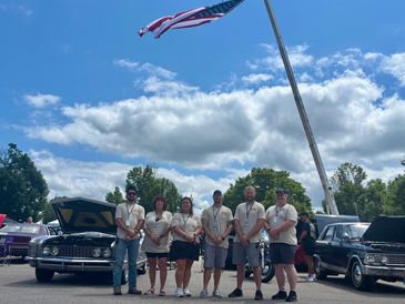 Six people stand in front of classic cars under a waving American flag on a sunny day.