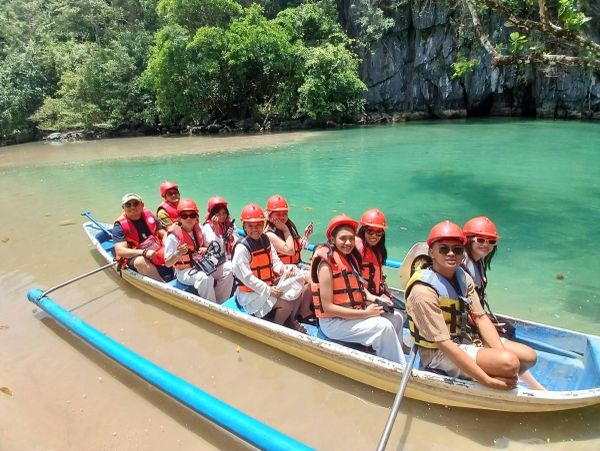 Group in a boat wearing life jackets and helmets by clear water.