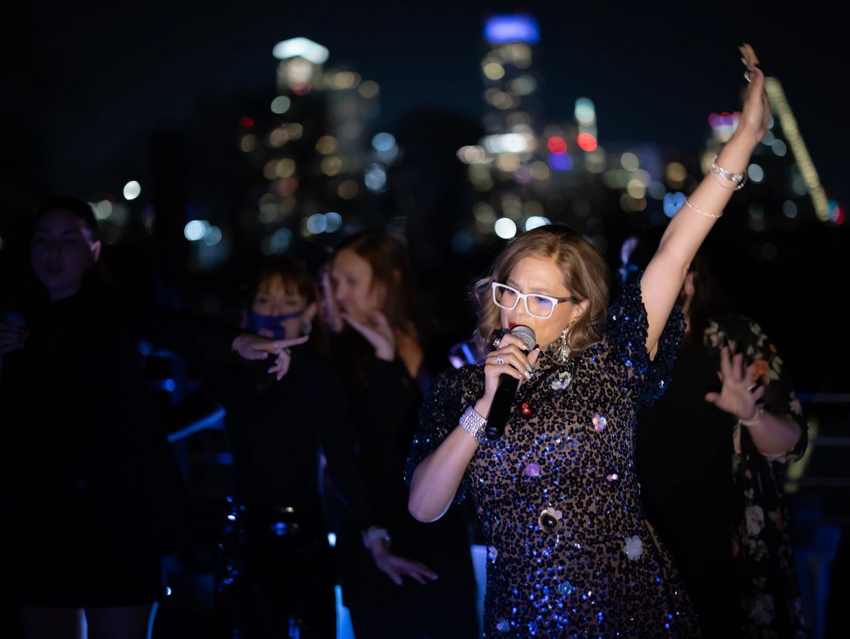 Woman singing on stage at night with city lights in the background.