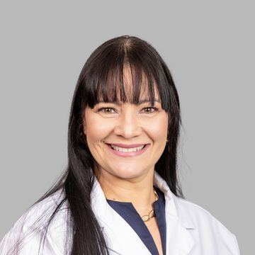 Smiling woman in a white medical coat with long dark hair and bangs.