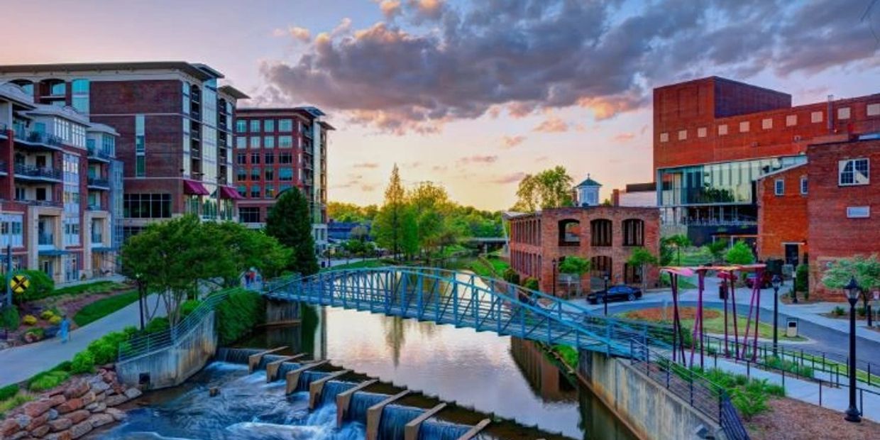Scenic urban riverwalk with modern buildings and a blue pedestrian bridge at sunset.