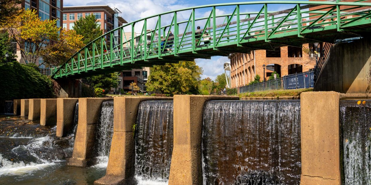 Green pedestrian bridge over a waterfall in an urban park setting.