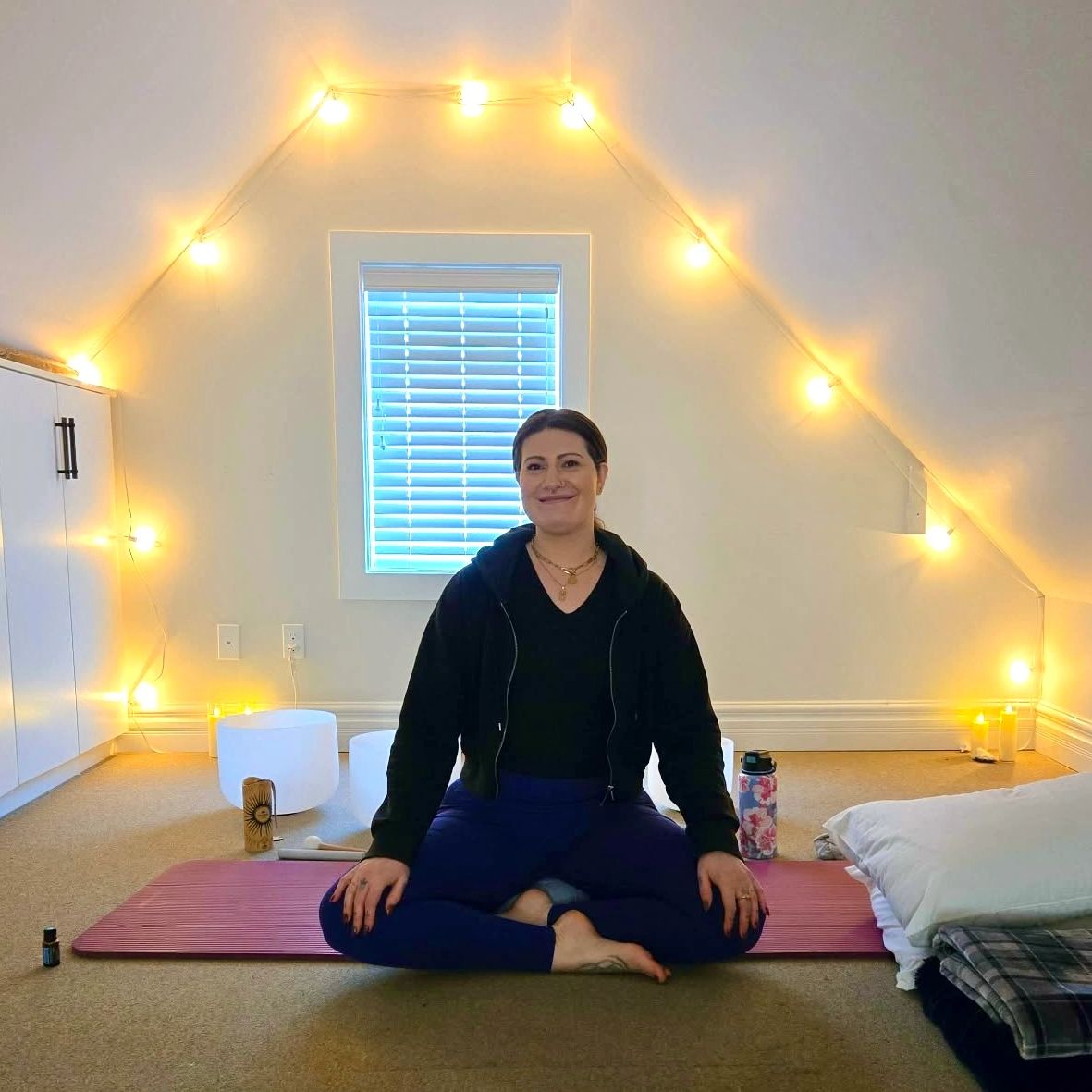 Woman sitting cross-legged on yoga mat in a cozy room with warm string lights.