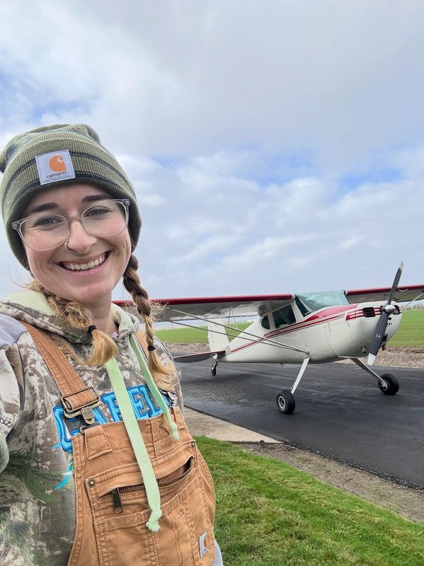 Smiling woman in outdoor gear stands near a small airplane on a runway.