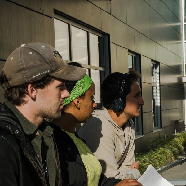 Three young adults attentively listening outdoors, one wearing headphones.