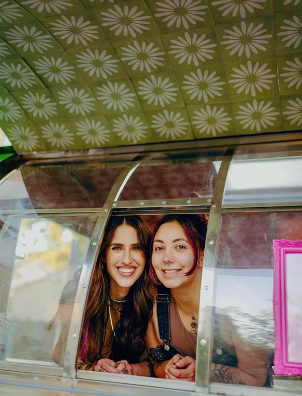 Two smiling women leaning out of a window with floral ceiling pattern above.