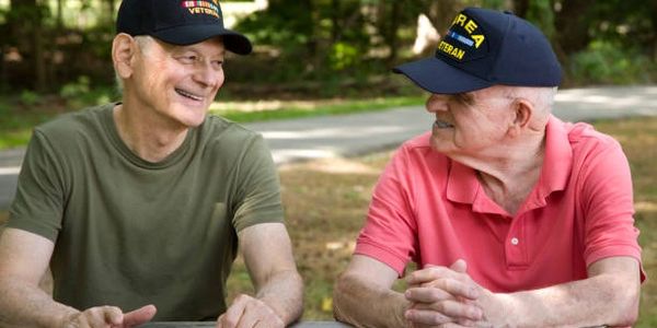 Two elderly veterans wearing caps, smiling at each other outdoors.