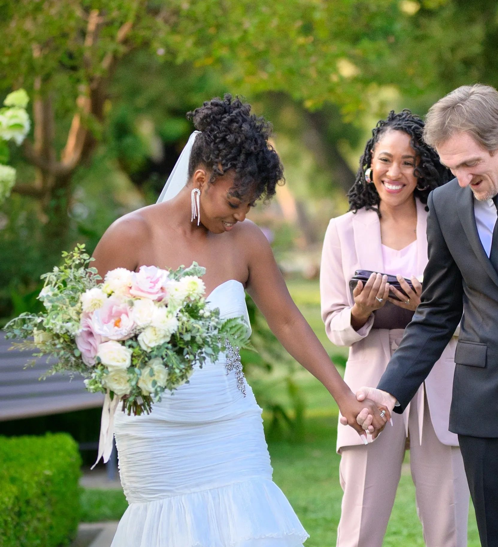 Bride and groom hold hands during outdoor wedding ceremony with officiant smiling behind.