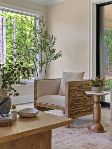 Cozy corner with a wooden armchair, side table, and greenery by large windows.