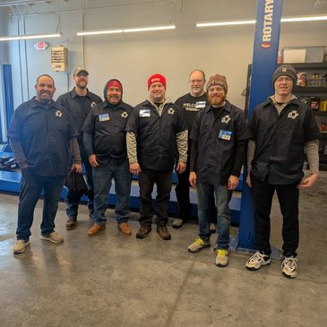 Group of seven men in work uniforms standing in a garage or workshop.