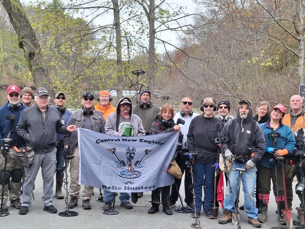 Group of metal detector enthusiasts posing outdoors with a banner.