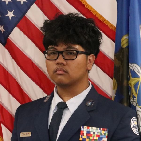 Young man in military uniform stands solemnly in front of American flag.