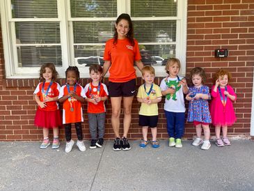 Young kids with medals stand beside their smiling teacher in front of a brick wall.