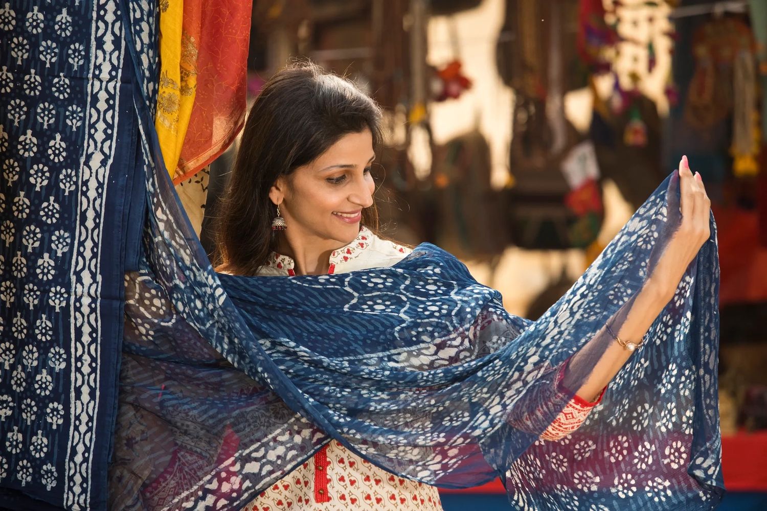 A woman admires a blue patterned scarf in a vibrant market setting.