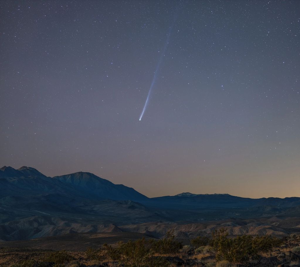 A bright comet streaks across a starry night sky above desert mountains.