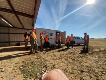 Group of hunters in orange vests gathered near a trailer on a sunny day.