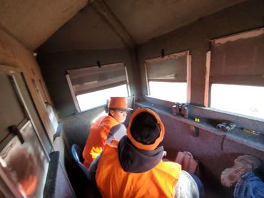 Two people in orange vests observing through windows inside a cabin.