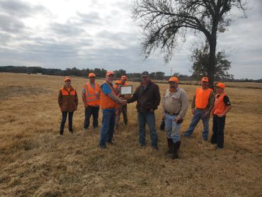 Group of hunters outdoors with a deer and a plaque, dressed in bright orange gear.