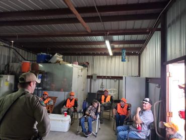 Group of people in orange safety vests sitting inside a metal shed.