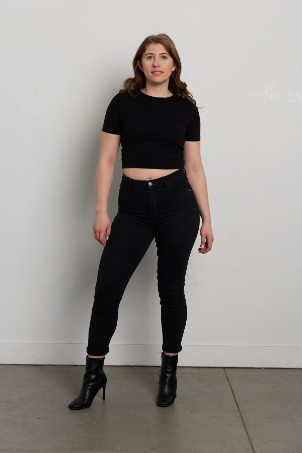 Woman in black crop top and jeans with ankle boots posing against white wall.