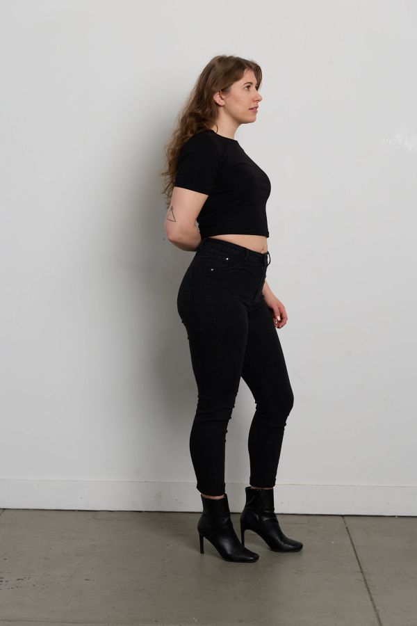 Woman in black outfit posing sideways against a plain wall.