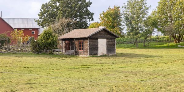 A small wooden cabin in a grassy field with trees and a red barn nearby.