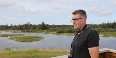 A man stands on a wooden deck overlooking a serene wetland landscape.