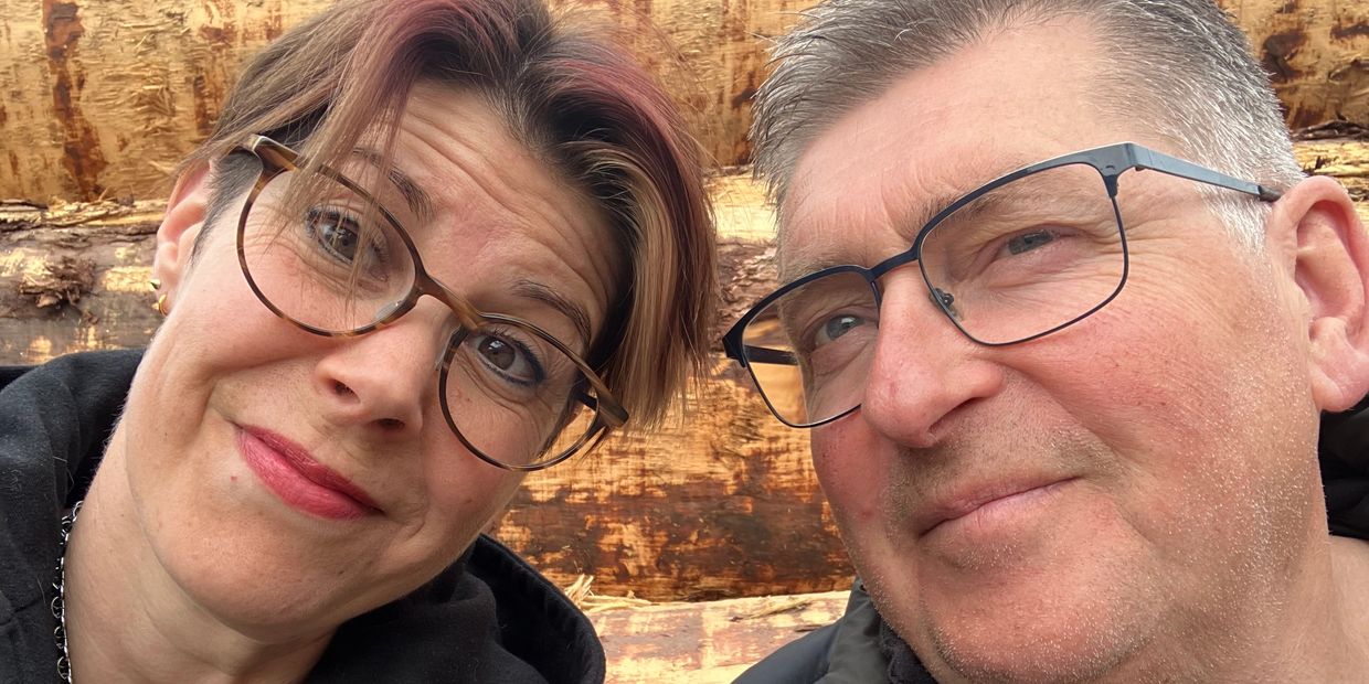 Close-up selfie of a smiling middle-aged couple wearing glasses outdoors.