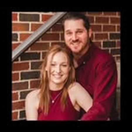 Couple posing happily in front of a brick wall, both wearing matching maroon tops.