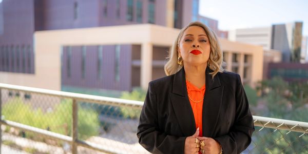 Confident woman in black blazer and red top stands outdoors in an urban setting.