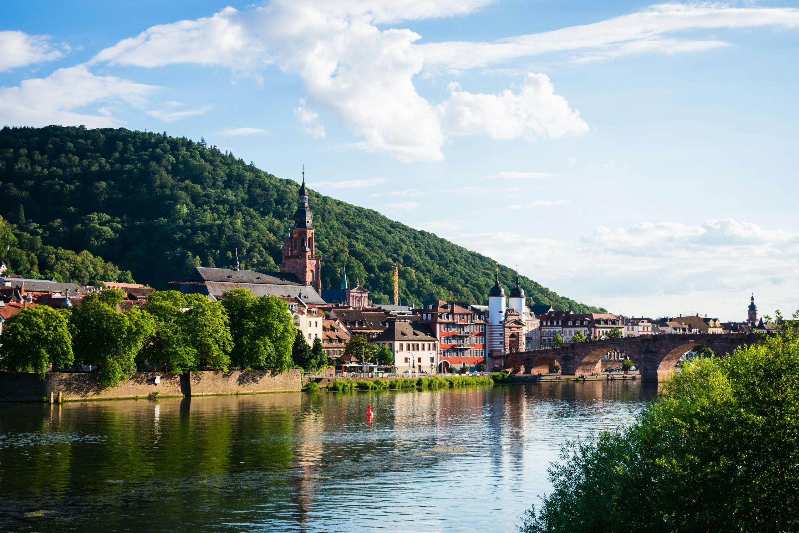 Scenic riverside town with historic buildings and a stone bridge under a blue sky.