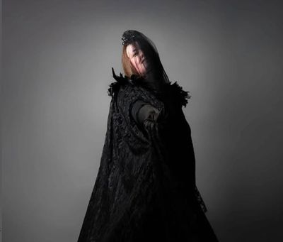 Person in a dramatic black cloak reaching forward against a gray background.