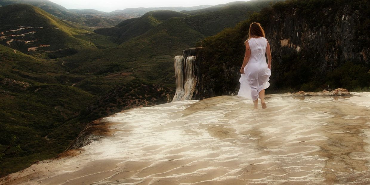 A woman in white walks near a scenic waterfall on a rocky ledge.