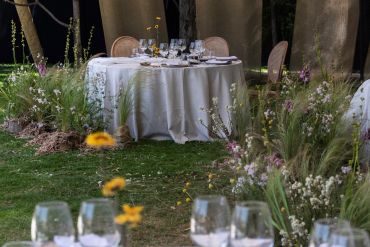 Mesa decorada para evento en jardín con flores y manteles blancos.
