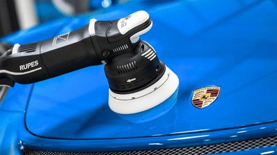 Polishing a blue Porsche car's hood with a Rupes tool.