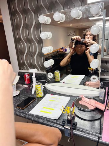 A woman sitting in front of a vanity mirror with a woman behind her working on her bangs