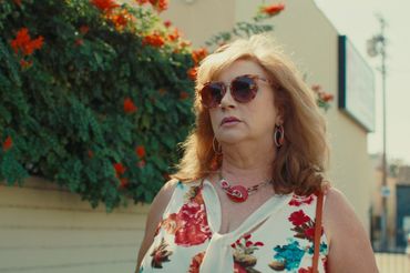 A woman wearing a floral top, big necklace and stylist shades posing in front of a rose bush outside