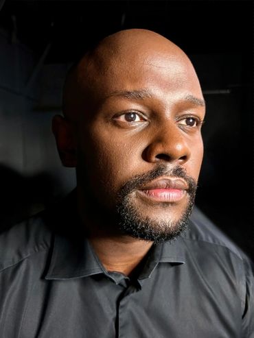 A dark skin man with a goatee wearing a black button up shirt staring off into the distance.