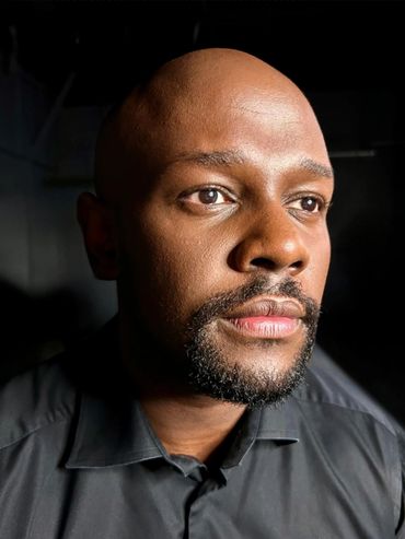 A dark skin man with a goatee wearing a black button up shirt staring off into the distance.