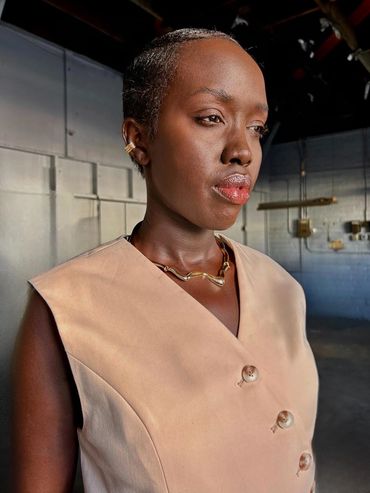 A dark skin woman with short hair wearing a tan vest and gold jewelry staring off into the distance.