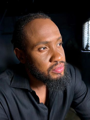 A dark skin man with a goatee wearing a black button up shirt staring off into the distance.