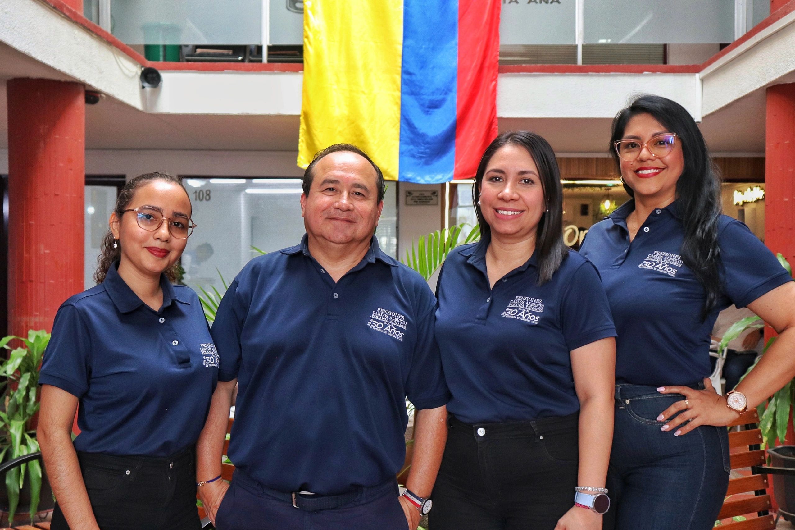 Four colleagues in matching navy blue polo shirts pose under a hanging Colombian flag.