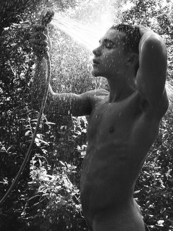A young man showers outdoors using a hose, surrounded by nature.