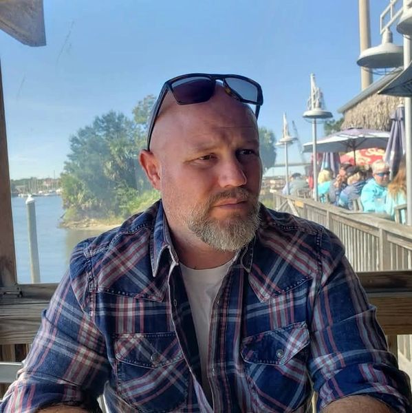 Man with sunglasses on head sitting outdoors near water.