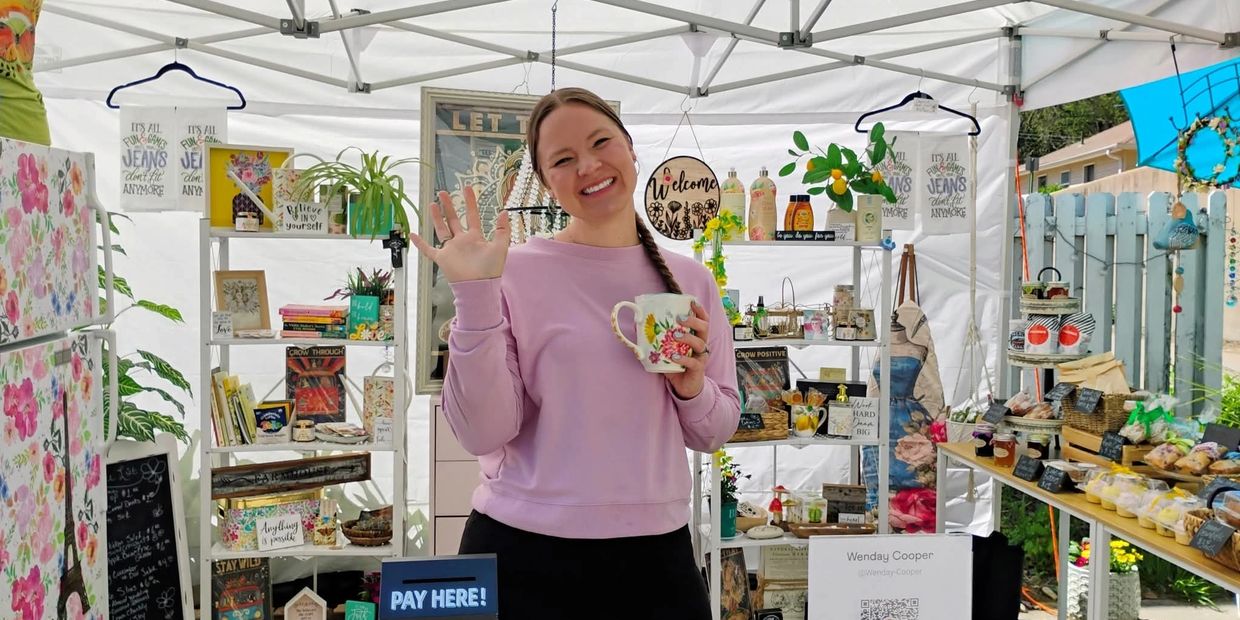 Smiling vendor waves while holding a floral mug at a colorful outdoor market stall.