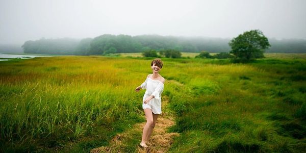A joyful woman in a white dress dances barefoot on a grassy path in a foggy meadow.