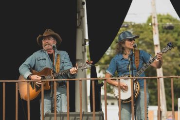 Two musicians playing guitar and banjo on stage outdoors.