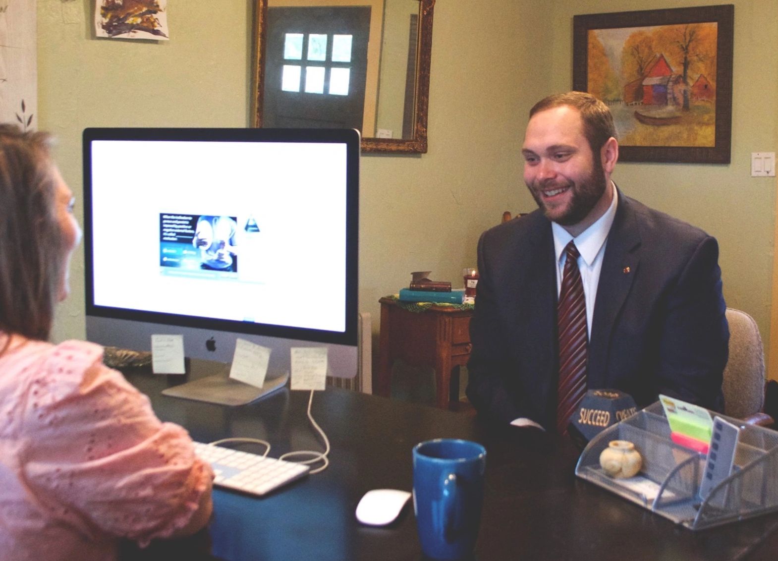 A man in a suit smiling during a meeting with a woman at a desk with a computer.